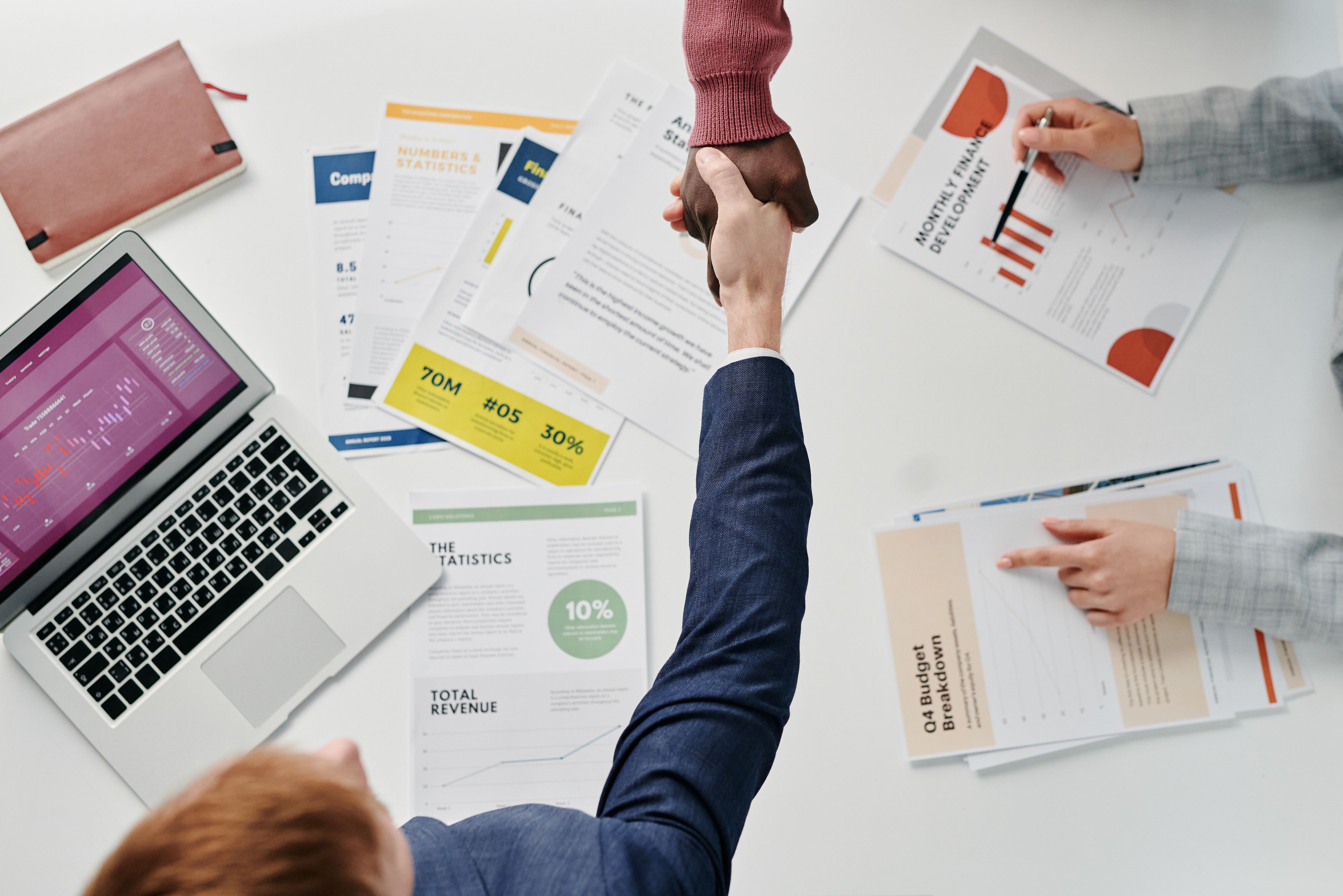 Business professionals engaging in a handshake over a desk filled with financial documents and a laptop