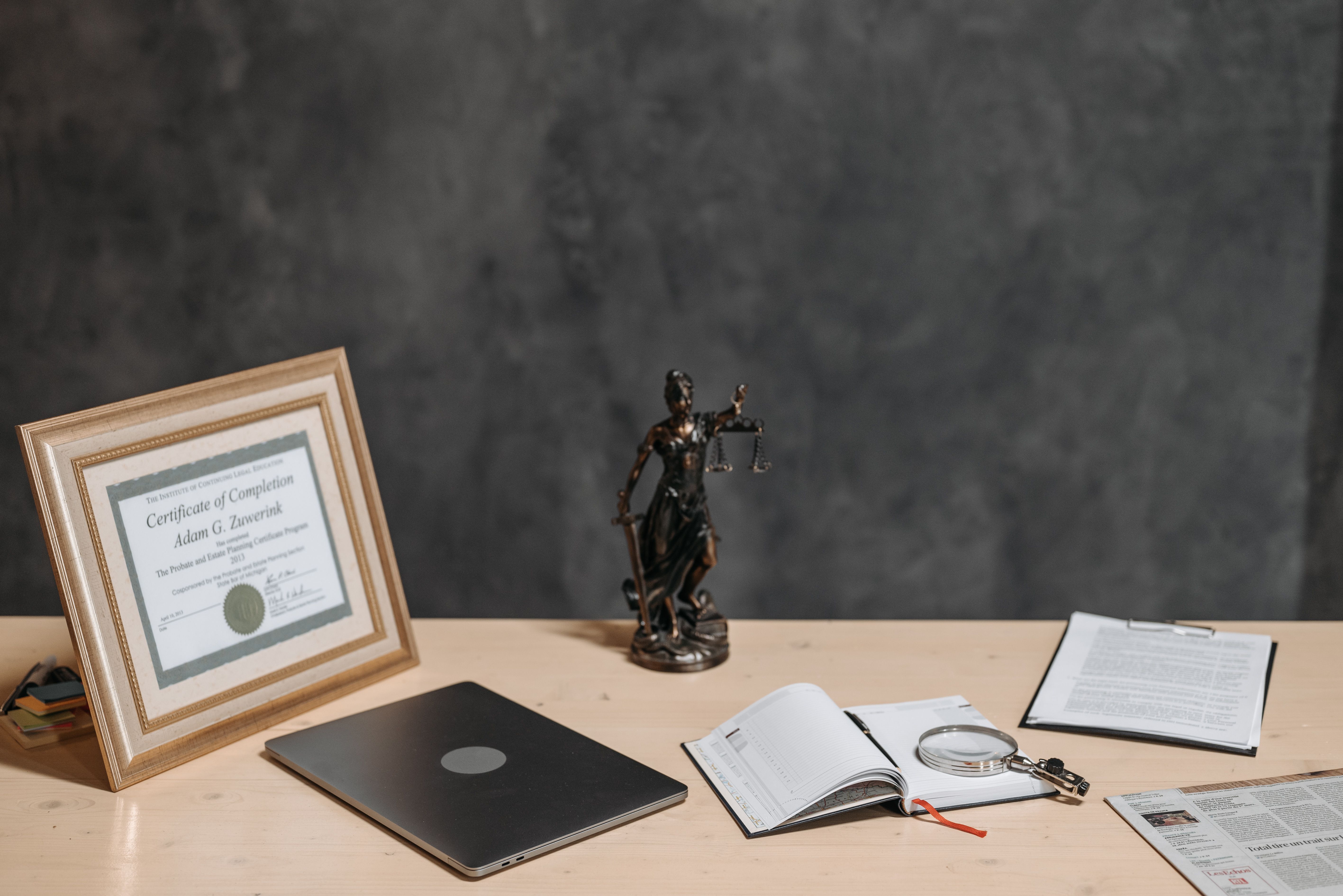 A lawyer's desk featuring a justice statue, diploma, laptop, magnifying glass, and documents
